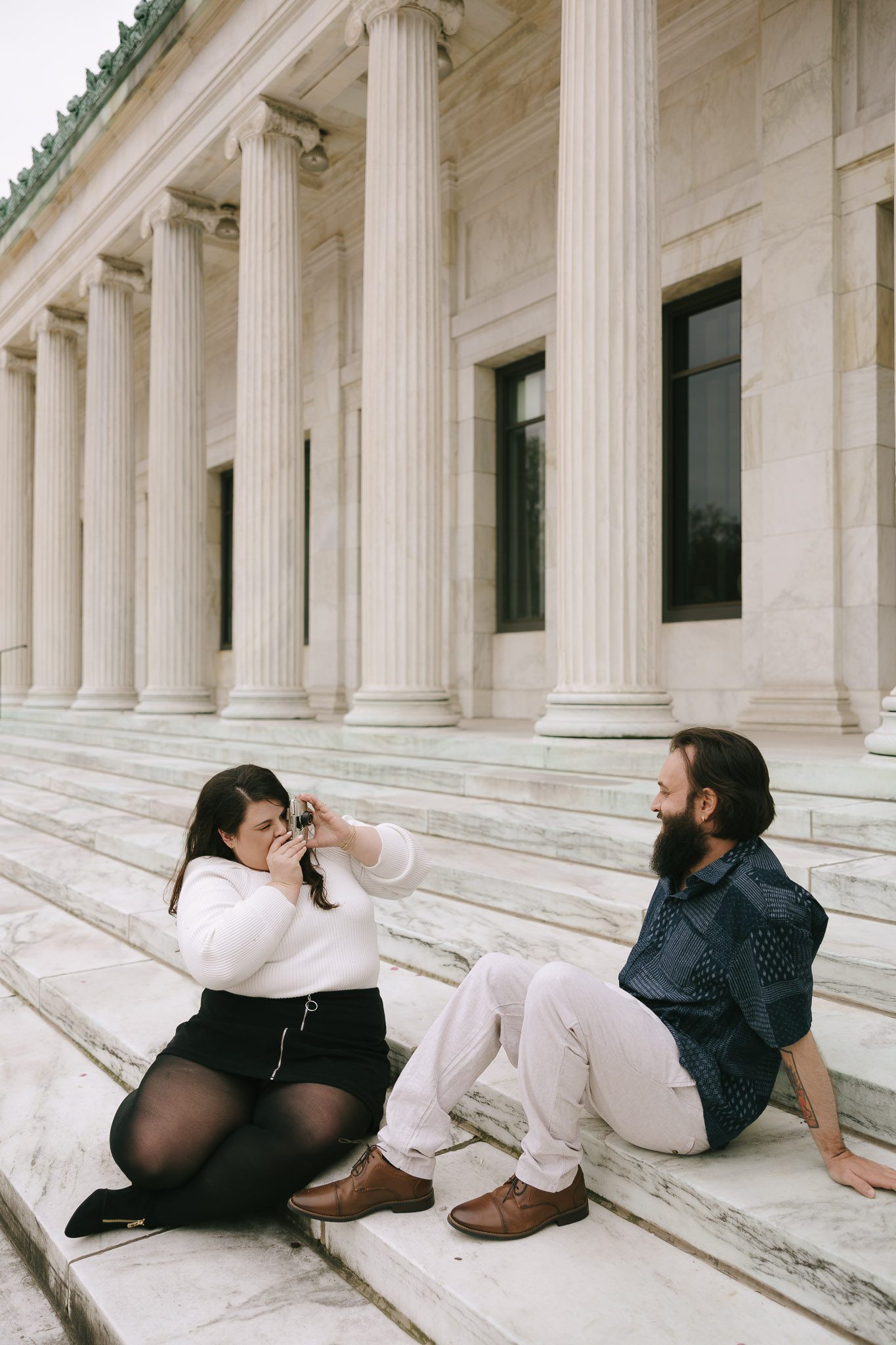 Bethany and Adam sit in front of the museum, taking a picture on a film camera