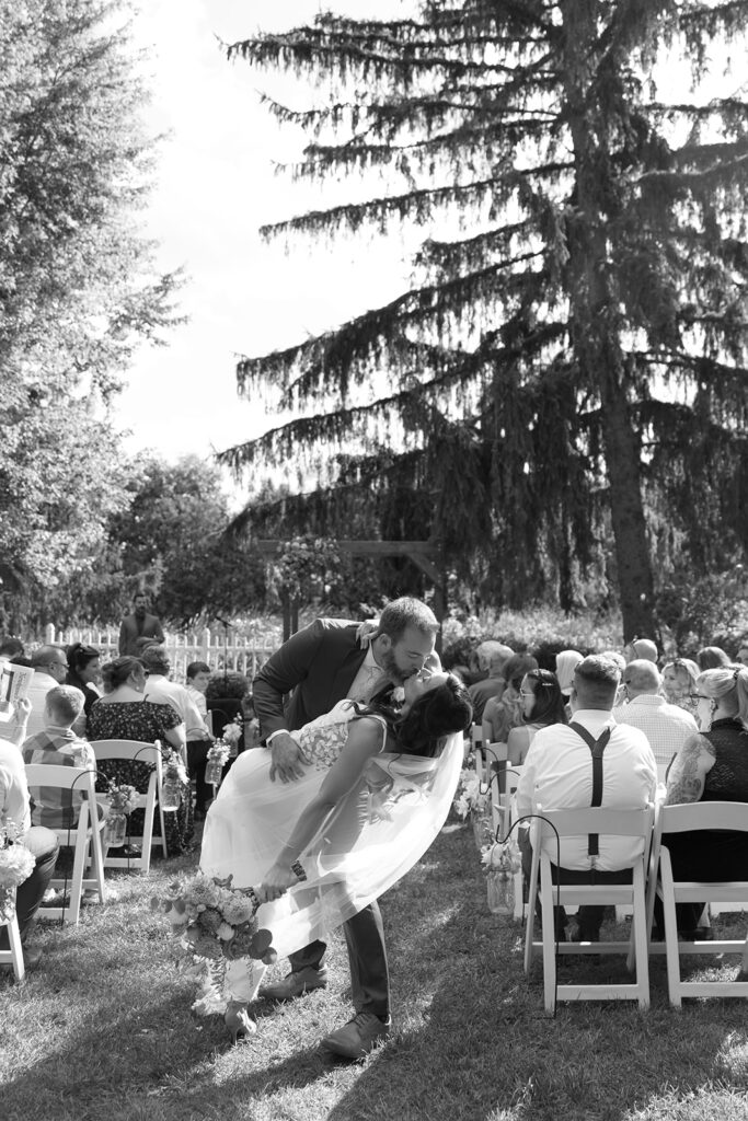 Tony dips Karis, as they walk back up the aisle during their wedding. Karis carries her wildflower bouquet and wears a custom dress, in black and white