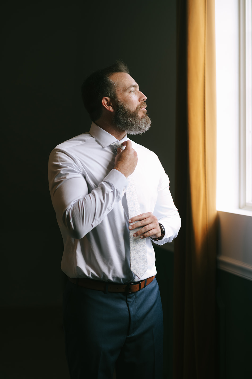 Tony tightens his tie while looking out the window at the grounds of Bloom and Bower