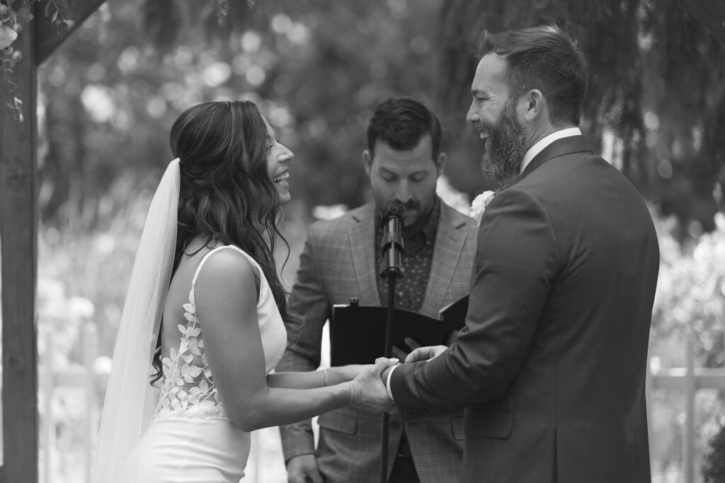 Karis and Tony laugh while exchanging rings under the wildflower arch at their bloom and bower wedding