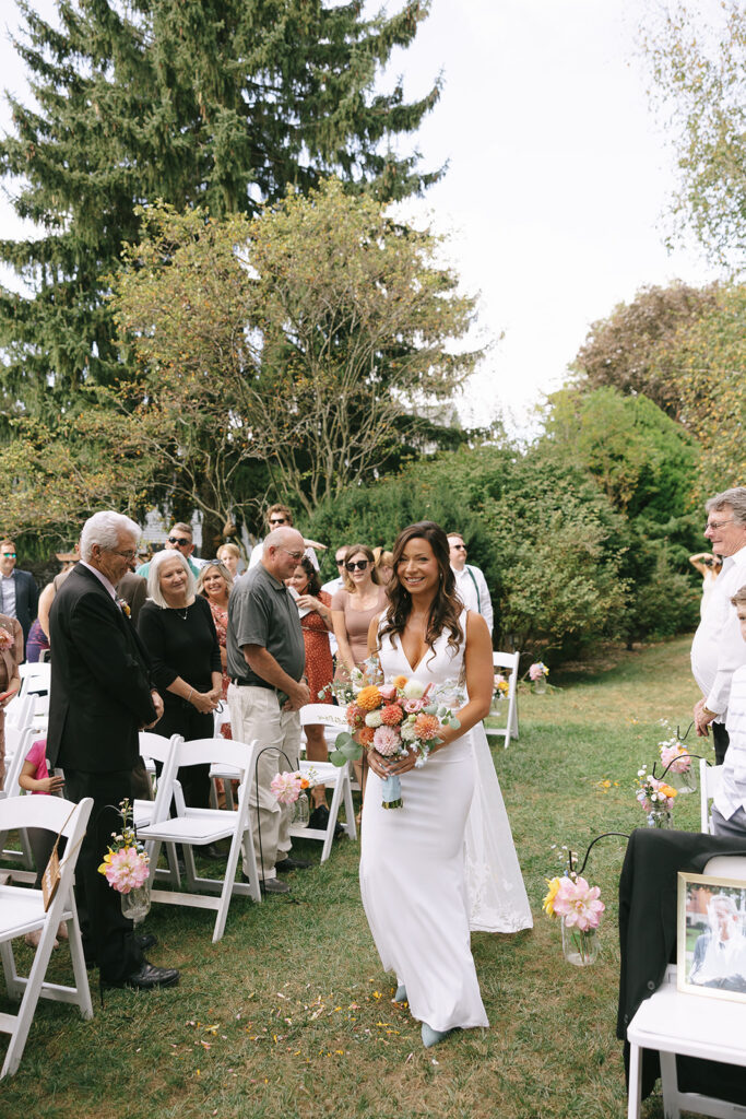 Karis walks down the aisle toward Tony on their wedding day at Bloom and Bower, carrying her wildflower bouqet and wearing a custom bridal gown