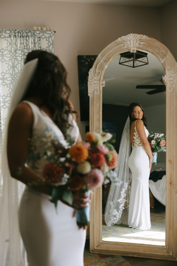 Karis peeks at her reflection in a full length mirror --seeing her custom dress with matching veil and wildflower bouquet