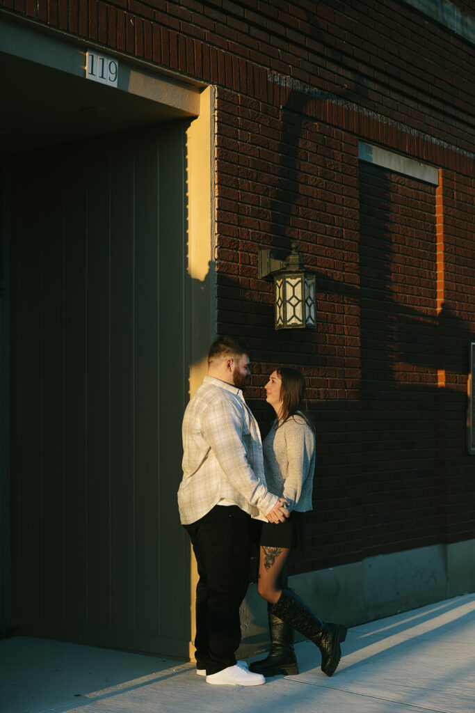 Vertical photo of a couple leaning close against a red brick wall, showing a natural and candid engagement moment in the city.