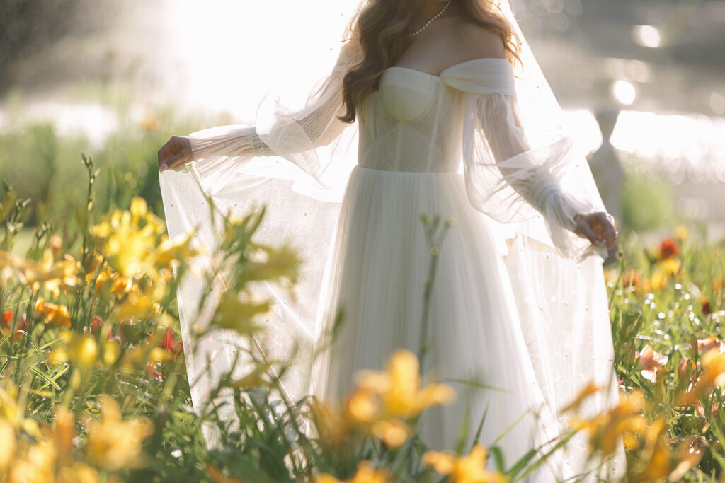 Bride in an off-the-shoulder tulle gown standing in a field of yellow flowers, holding her skirt gently as sunlight glows behind her. Blog title of define Your Bridal Look