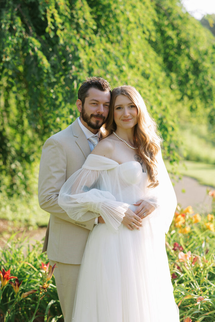 Groom in a beige suit holding his bride from behind as they smile softly, framed by lush greenery and colorful flowers. Blog title of define Your Bridal Look