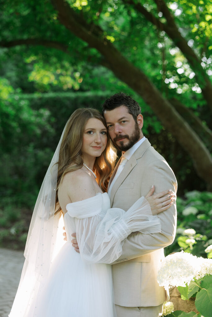 Bride and groom standing close together in a shaded garden, gazing into the camera with a quiet, intimate expression.