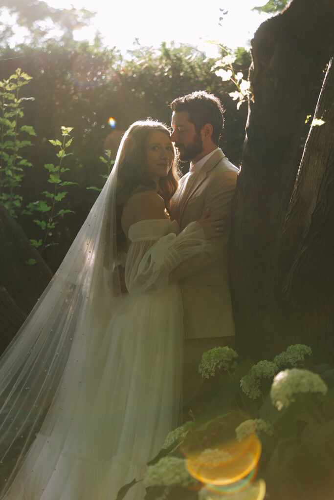Bride and groom standing close in a shady garden, sunlight filtering through trees and creating a warm, romantic glow.