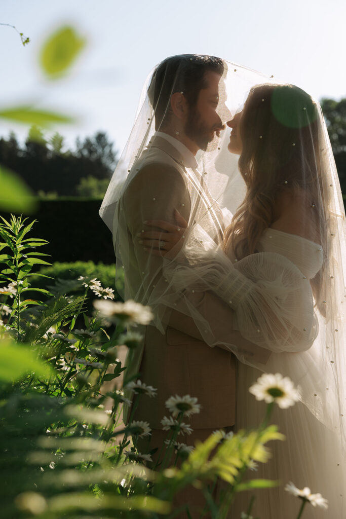 Bride and groom wrapped in the bride’s pearl-dotted veil, facing each other with their noses almost touching in soft evening light.


