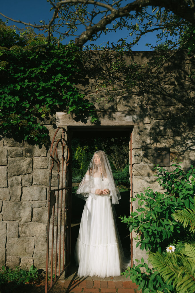 Bride standing in a stone archway, veil draped over her face, eyes closed as golden sunlight illuminates her gown and surroundings.