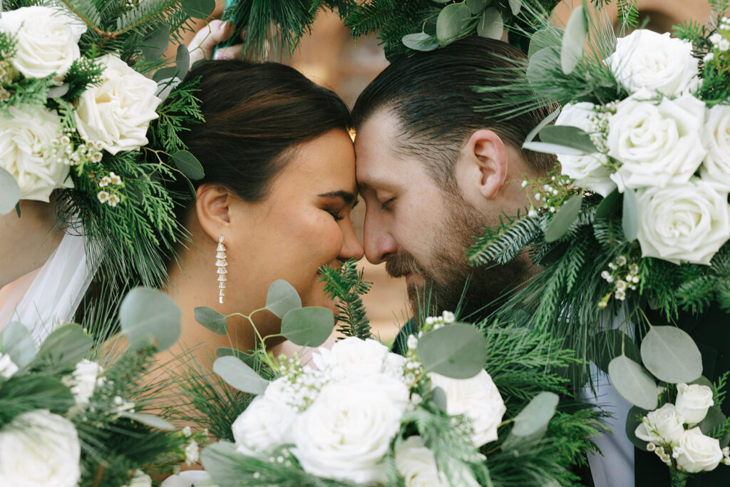 Bride and groom framed by bouquets of white roses and greenery, showing the connection couples look for when finding a wedding photographer on Instagram.