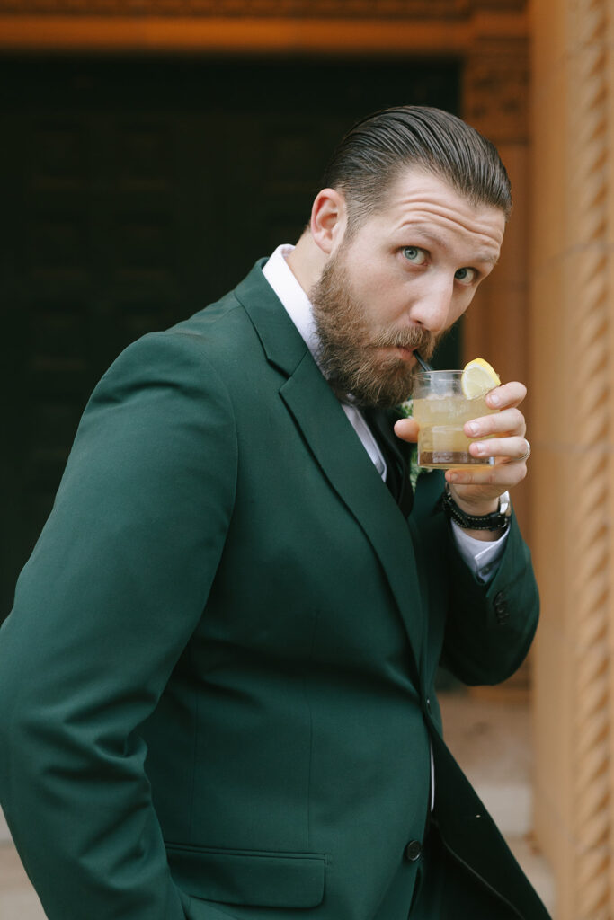 Groom in dark green suit sipping a cocktail outside a brick venue, personality-driven photo style shown when finding a wedding photographer on Instagram.
