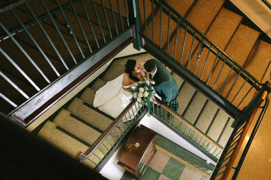Overhead shot of bride and groom kissing on a staircase inside a historic building, creative angle couples often notice when finding a wedding photographer on Instagram.