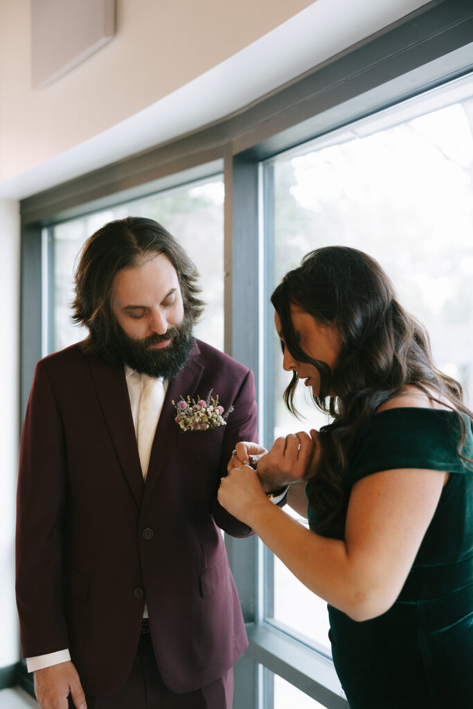 Groom in burgundy suit getting boutonniere pinned with natural window light, an example of good prep lighting in tips for the morning of your wedding.