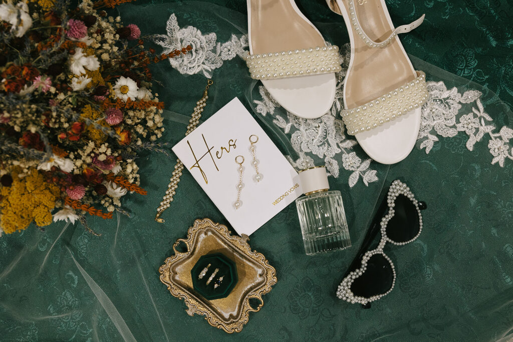 Flat lay of bridal details including bouquet, pearl shoes, vow book, perfume, and jewelry styled for photos — a must-have in tips for the morning of your wedding.