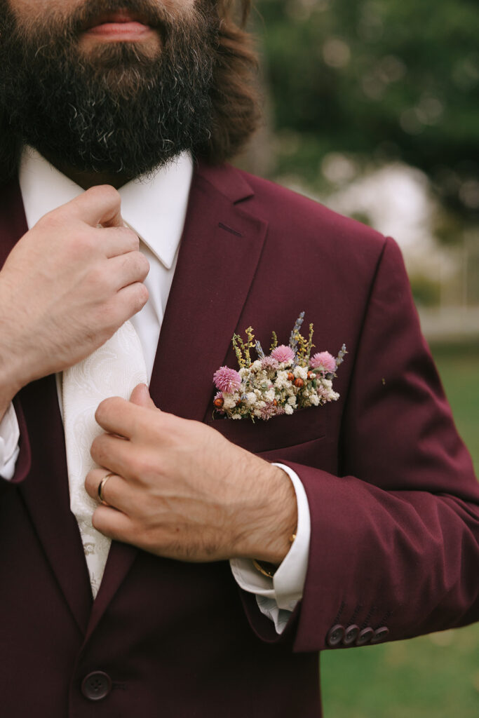Close-up of groom adjusting burgundy suit jacket with boutonniere, natural portrait for tips for the morning of your wedding.