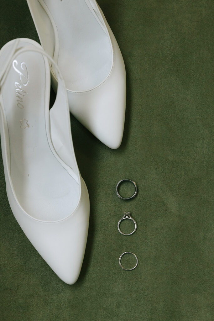 wedding day timeline details featuring bridal shoes, engagement ring, wedding bands, and invitation in a flat lay