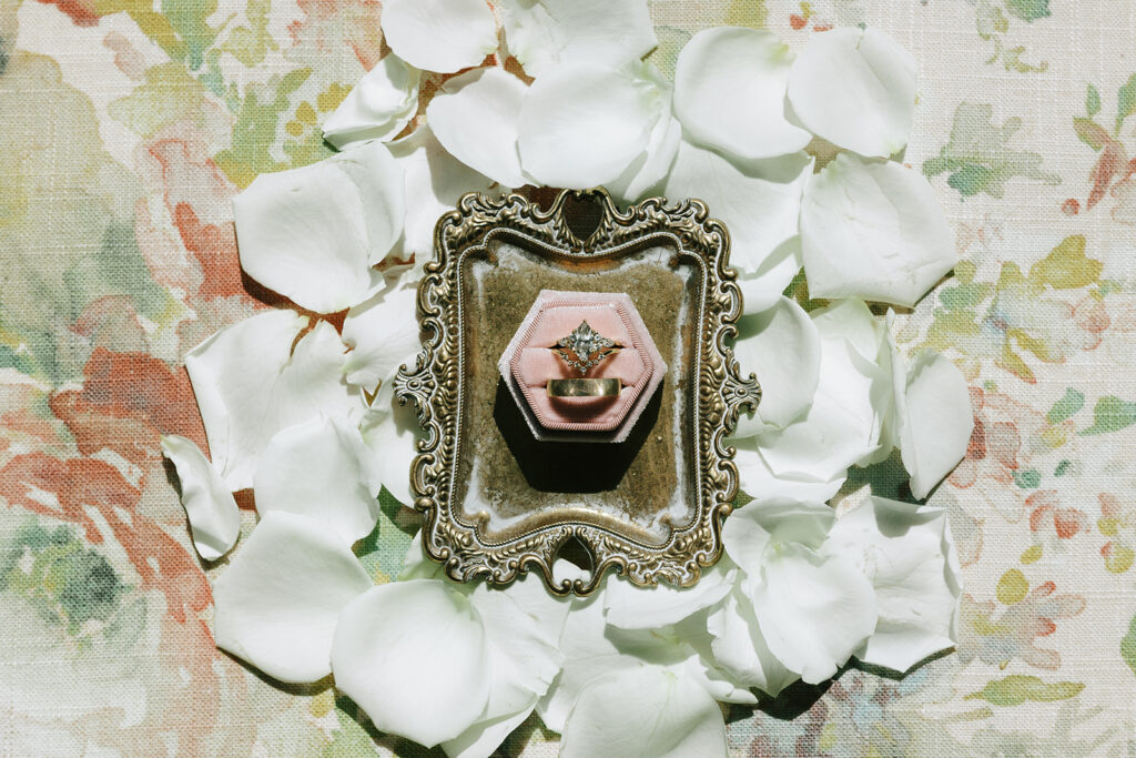 A vintage tray holding a pink ring box with an engagement ring and wedding band, surrounded by white rose petals on a floral fabric background. A styled detail photo showing accessories frequently found in Black Friday wedding deals.

best Black Friday wedding deals