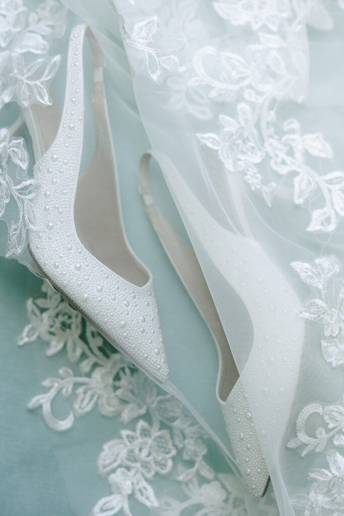 A pair of pearl-embellished white bridal heels resting on embroidered lace fabric. A wedding shoe detail photograph featuring items couples often look for during Black Friday wedding deals.

best Black Friday wedding deals