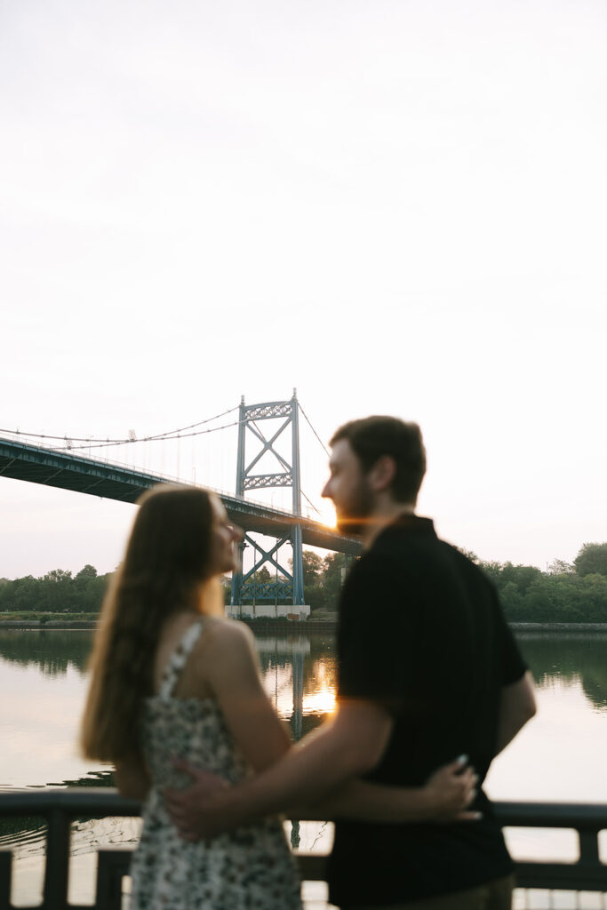 A couple stands close together by the river as the sun sets behind the blue suspension bridge, creating a warm glow through the frame. This emotion-driven moment is often used to support small business photography portfolios.