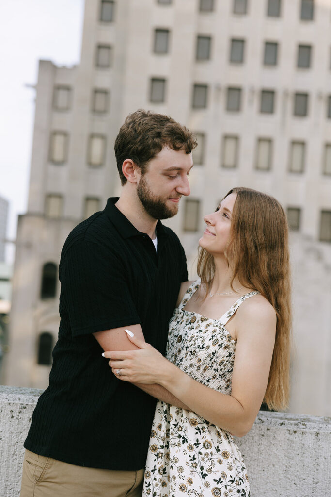 A couple embraces on a rooftop ledge downtown, looking into each other’s eyes with soft smiles. A natural, heartfelt moment that supports small business photographers showcasing genuine connection.