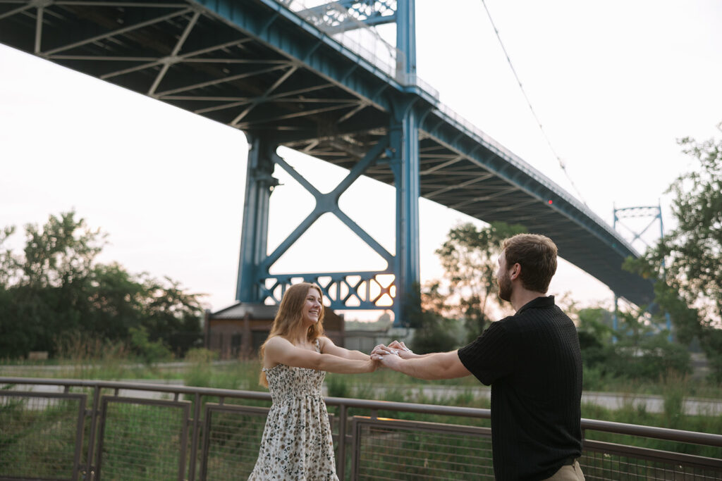 A couple smiles at each other while holding hands on a riverside walkway beneath a tall blue suspension bridge at sunset. The warm, candid moment highlights connection and community an image often used to support small business storytelling.