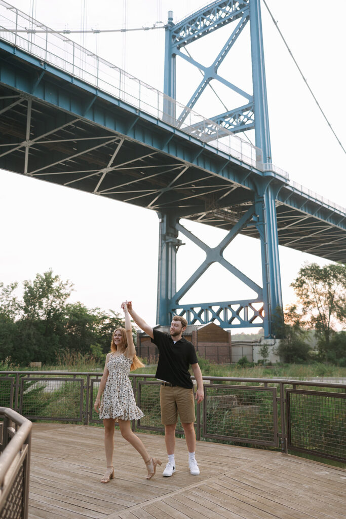 A couple dances on a wooden platform beneath a towering blue bridge, spinning with relaxed joy during a sunset session. The scene reflects natural moments that support small business creatives.
