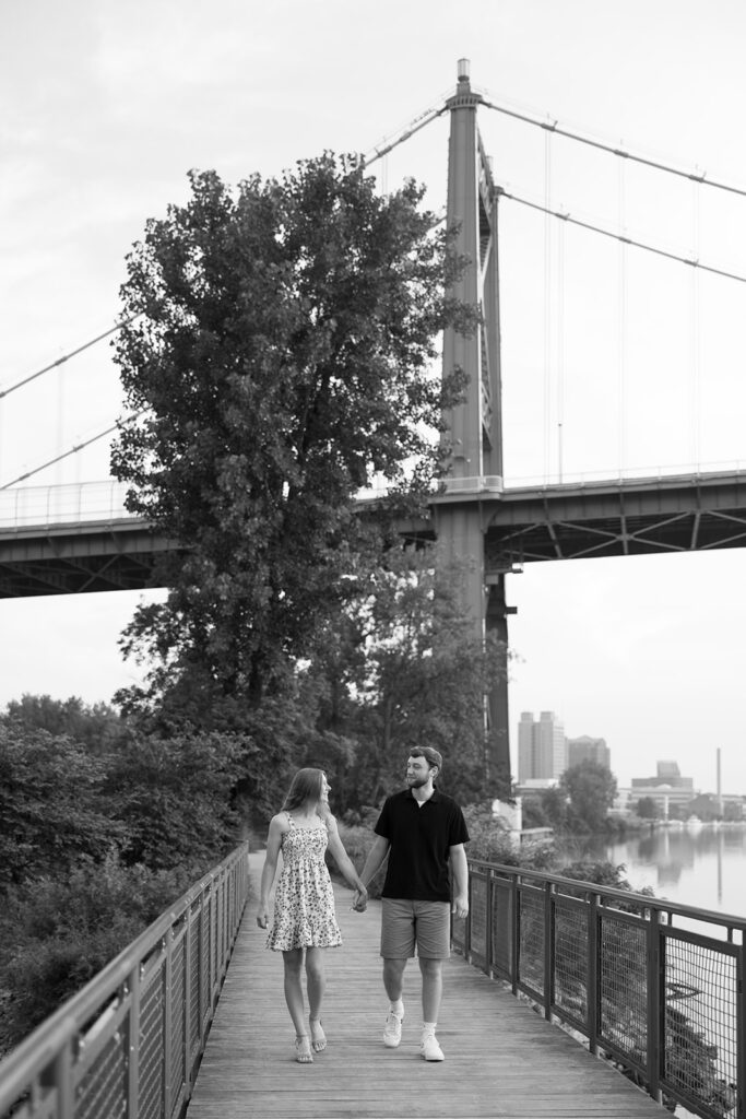 A couple walks hand-in-hand along a tree-lined river path beneath a tall bridge in a black-and-white photograph. The candid feel supports small business creatives showcasing authentic connection.