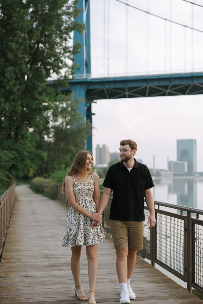A couple walks together along a riverside boardwalk, holding hands and smiling as the city skyline glows softly behind them. A natural moment that helps support small business photography content.