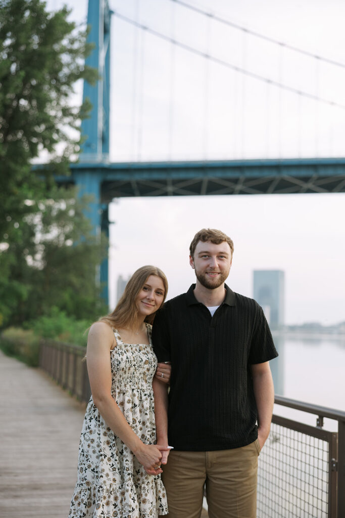 A couple stands arm-in-arm on the riverside boardwalk with the skyline behind them, looking relaxed and comfortable together. This warm portrait supports small business creatives who capture everyday love.
