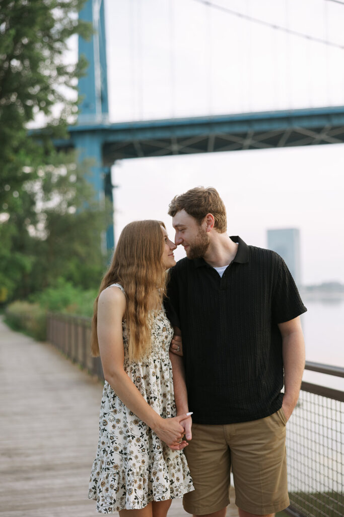 A couple stands close together on a wooden riverside path, leaning in forehead to forehead under a large blue bridge. The soft evening light adds a romantic feel perfect for posts that support small business photography.