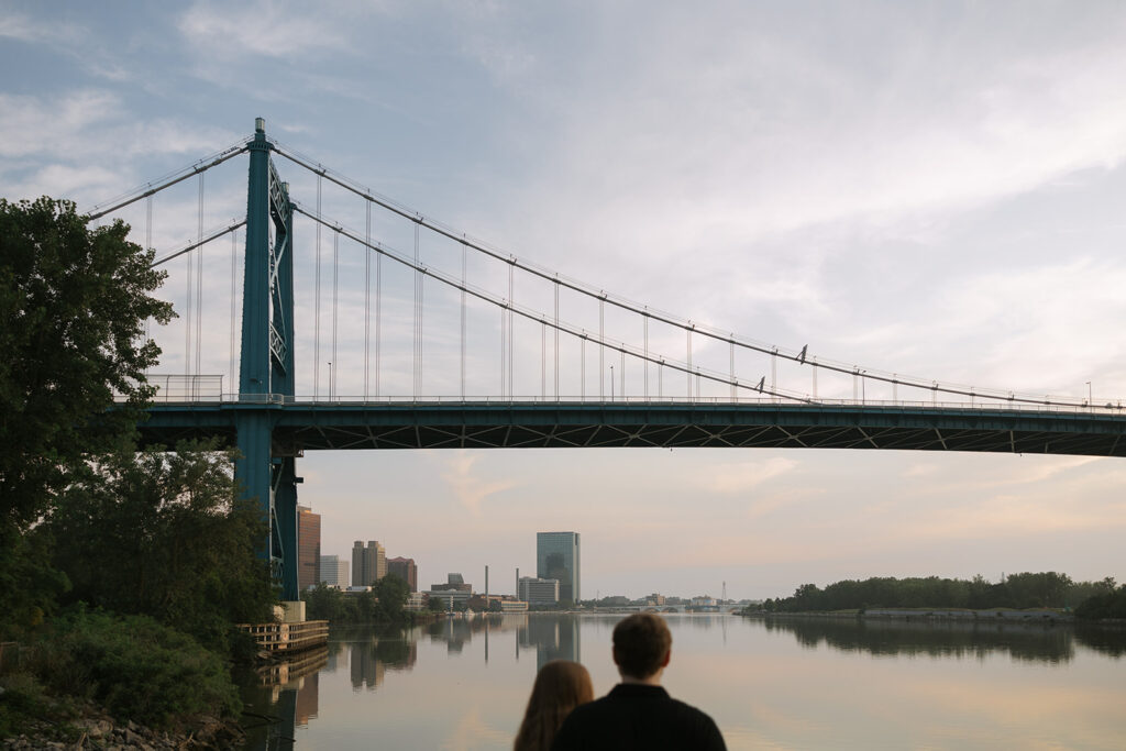 A couple stands with their backs to the camera as they look out over a calm river and distant city skyline beneath a long blue suspension bridge. The peaceful scene captures the kind of storytelling imagery that helps support small business photographers.