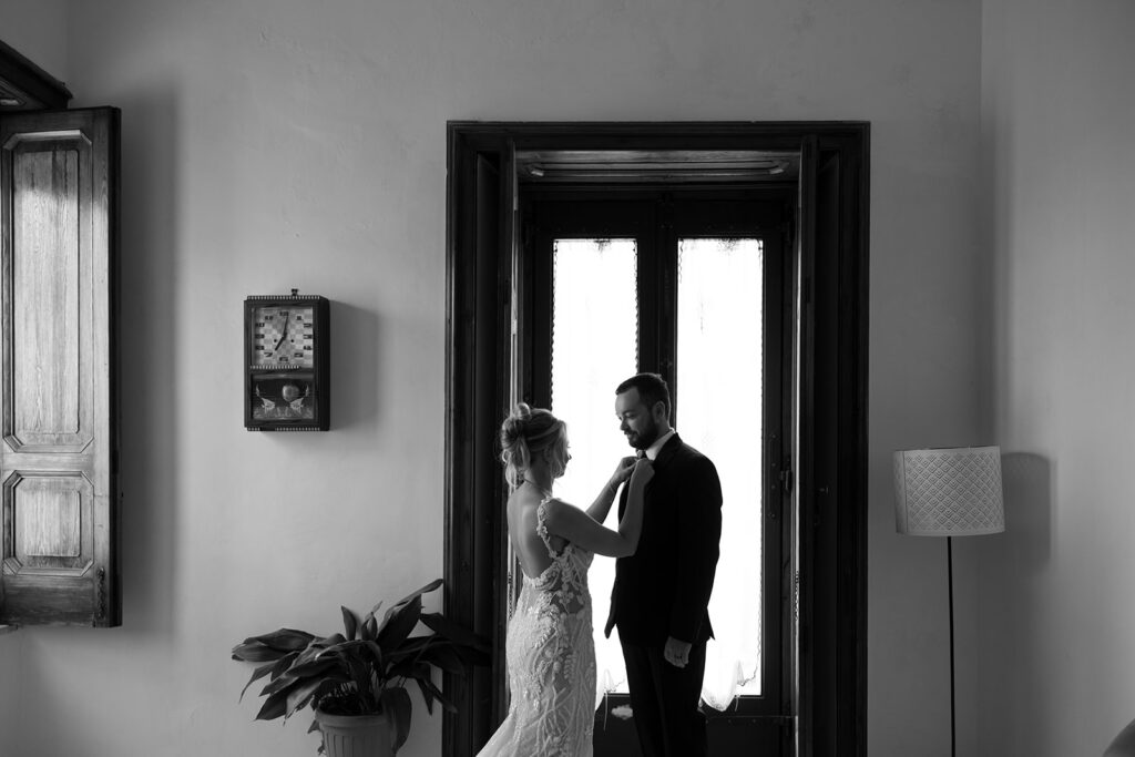 Black and white image of woman and man facing each other with the woman touching the man's tie. They are in a room with a clock on the wall, a door behind them, and a lamp on the floor.