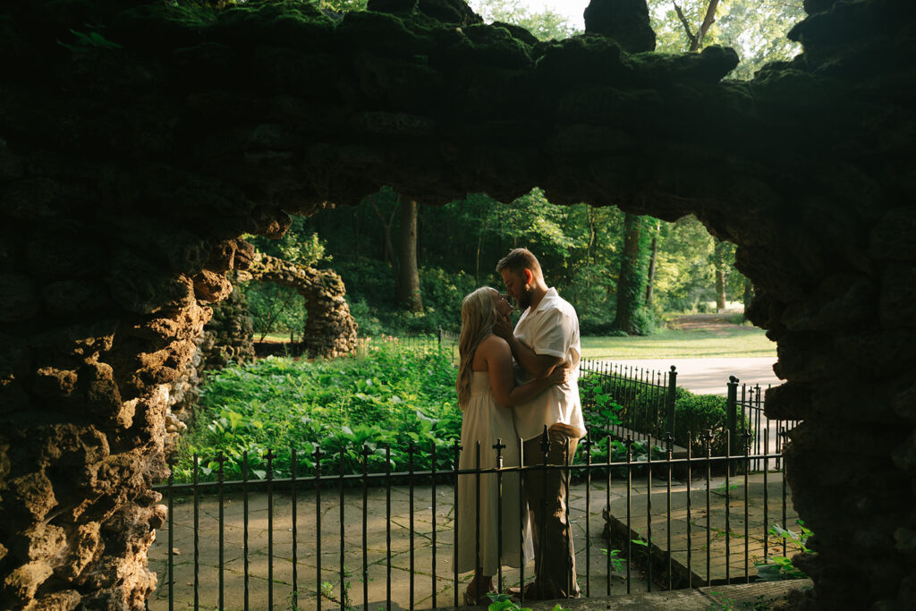 Couple standing close together beneath a rustic stone archway in a lush green park during an anniversary photo session, framed by soft natural light.
