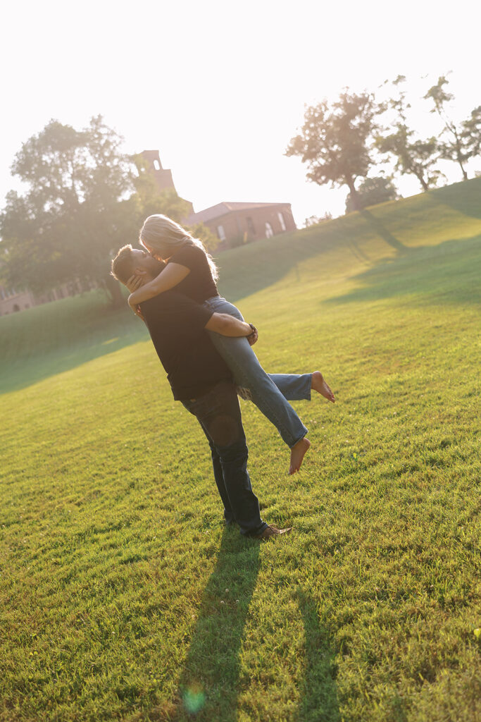 Engaged couple laughing as one partner lifts the other during a golden hour engagement session on a grassy hill 