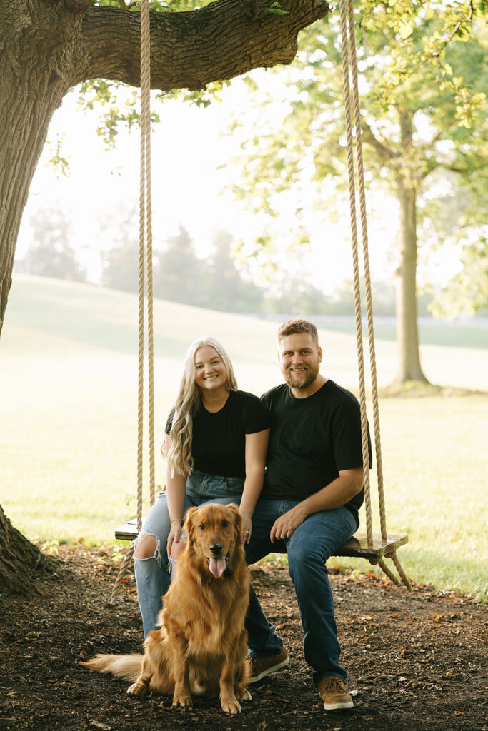 Engaged couple sitting on a wooden tree swing with their golden retriever during an outdoor engagement session 