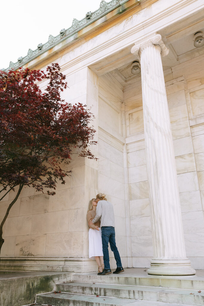 A man and a woman next to a column, kissing, for their engagement photos.