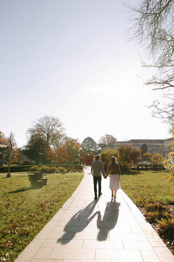 A man and a woman walking away from the calera, holding hands with long shadows behind them for their engagement photos.
