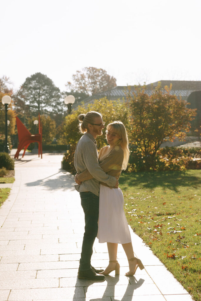 A man and a woman hugging around the waist, standing on a sidewalk for their engagement photos.