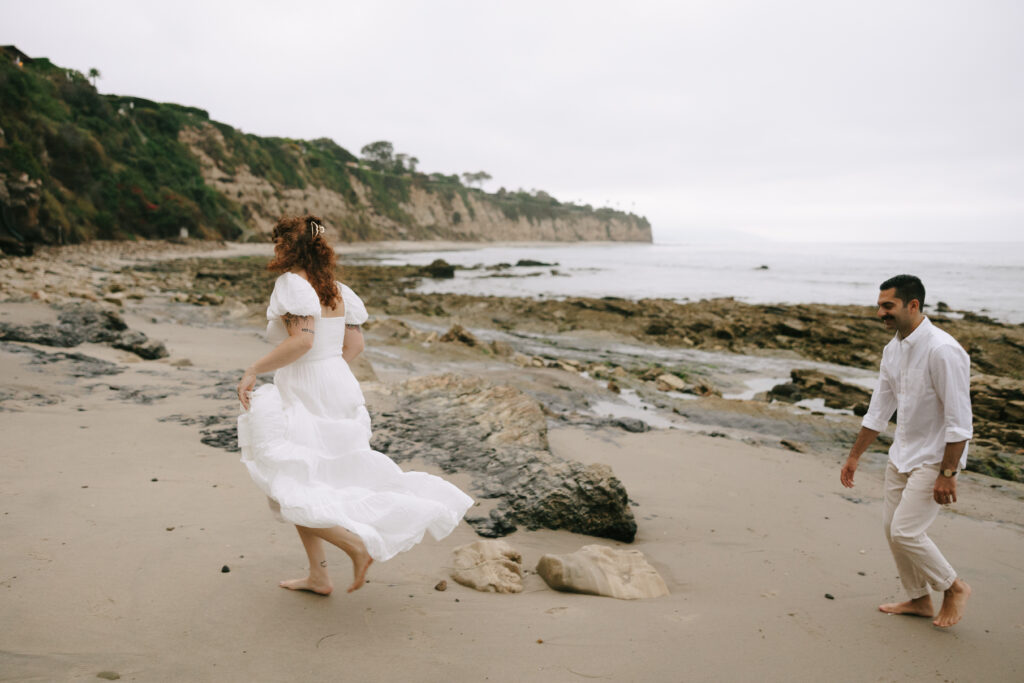 Toledo waterfront wedding portraits featuring the beach with a woman in white and a man walking behind her in white and cream. 