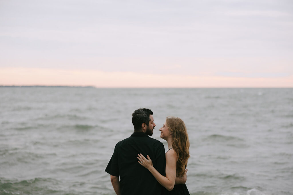 Woman and man on the beach smiling and holding each other with the water in the background.